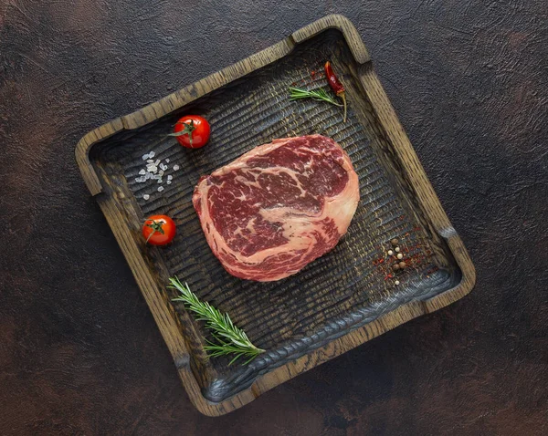 Raw meat Black Angus for Rib eye or Striploin steak on wooden cutting plate with fresh rosemary,cherry tomatoes and dried papper.Top view.