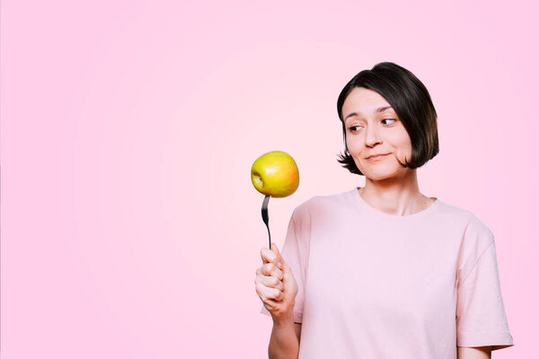 Young Woman holds in hand a fork with green apple on a light pink background. Healthy organic food for dieting woman concept