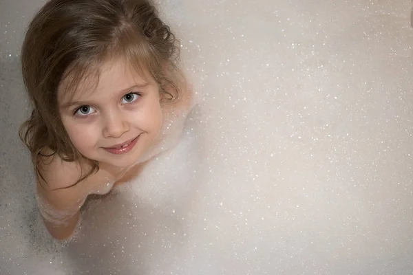 Щаслива усміхнена маленька дівчинка сидить у ванній з повною піною — стокове фото