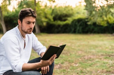 Parkta oturan genç adam dikkatle eski bir kitabı okur..