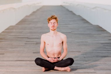 Genç kızıl saçlı adam nilüfer yogası yapıyor, Padmasana, açık havada, ahşap zeminde.