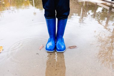 Mavi lastik çizmeli bir çocuk parktaki su birikintisinin üzerinden atlıyor..