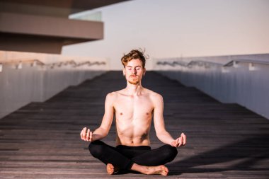 Genç bir yogi, nilüfer çiçeğinde tahta üzerinde otururken ellerini dizlerinin üzerine koyar. Boşluğu kopyala