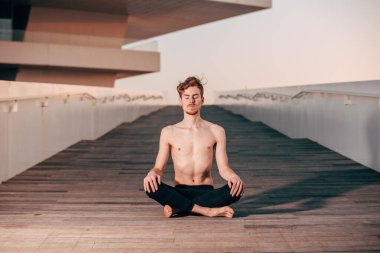Genç yoga pratisyeni poz veriyor, bacak bacak bacak üstüne atıyor alacakaranlıkta şehrin ahşap zemininde oturuyor, lotus poz veriyor..