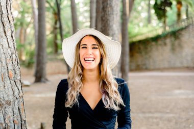 Büyüleyici genç manken parkta hasır şapkasına dokunurken mutlu bir şekilde gülüyor..