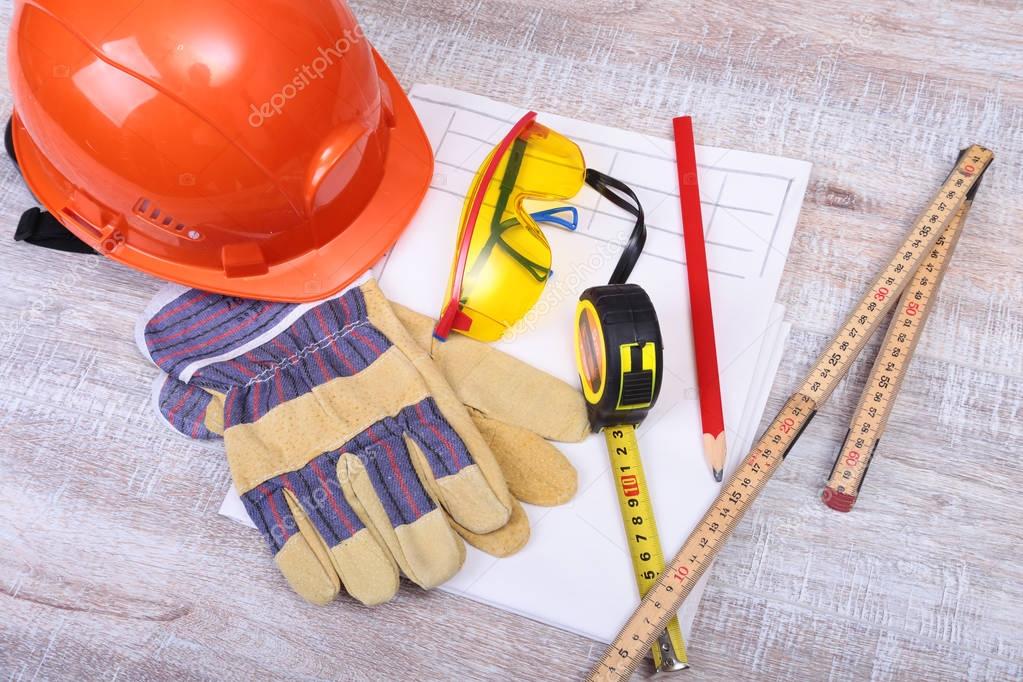 Orange hard hat, safety glasses, gloves, pen and measuring tape on wooden background.