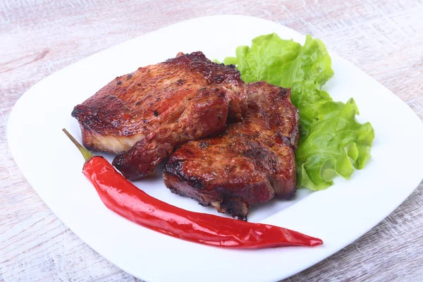 Steak of grilled meat and chili pepper with tomato, lettuce leaves on white plate.