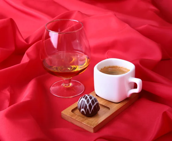 White cup of coffee and cognac in a glasses, pralines on red background. — Stock Photo, Image