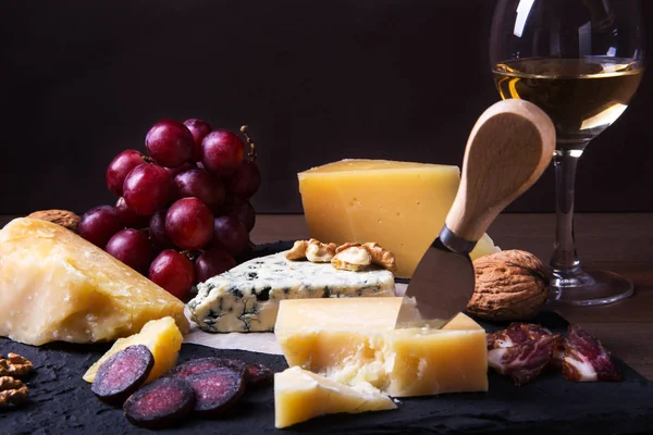 Quesos variados, frutos secos, uvas, frutas, carne ahumada y una copa de vino en una mesa de servir. Estilo oscuro y Moody. Espacio libre para texto . — Foto de Stock