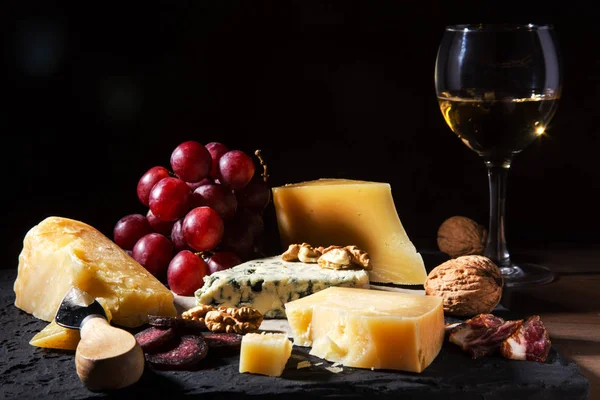 Quesos variados, frutos secos, uvas, frutas, carne ahumada y una copa de vino en una mesa de servir. Estilo oscuro y Moody. Espacio libre para texto . — Foto de Stock