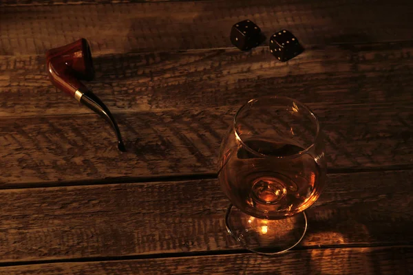 Szkło z koniakiem lub whiskey i fajka do palenia na drewnianym stole. Mroczny i humorzasty styl. — Zdjęcie stockowe