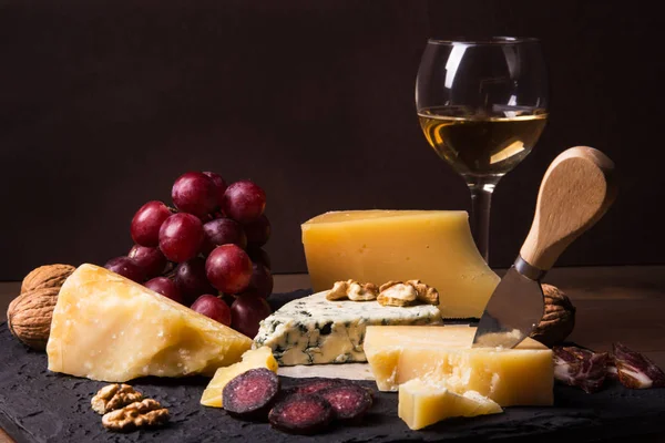 다양한 치즈, 견과류, 포도, 과일, 훈제 고기, 와인 한 잔이 서빙 테이블에 있습니다. 어둡고 무디 스타일. 텍스트의 여유 공간. — 스톡 사진