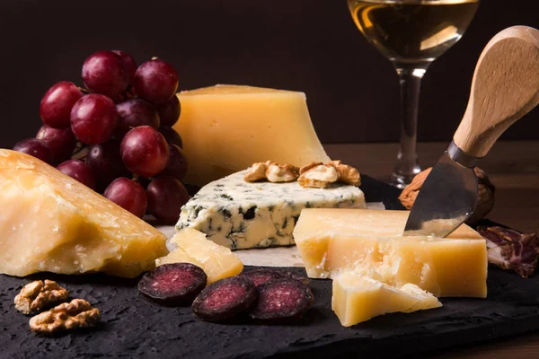Diverse ostar, nötter, vindruvor, frukter, rökt kött och ett glas vin på ett serveringsbord. Mörk och lynnig stil. Ledigt utrymme för text. — Stockfoto