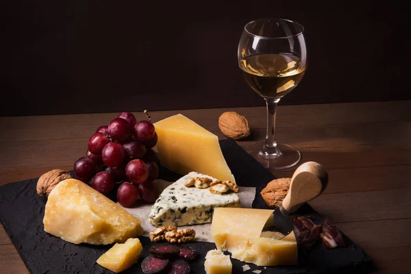 Diverse ostar, nötter, vindruvor, frukter, rökt kött och ett glas vin på ett serveringsbord. Mörk och lynnig stil. Ledigt utrymme för text. — Stockfoto