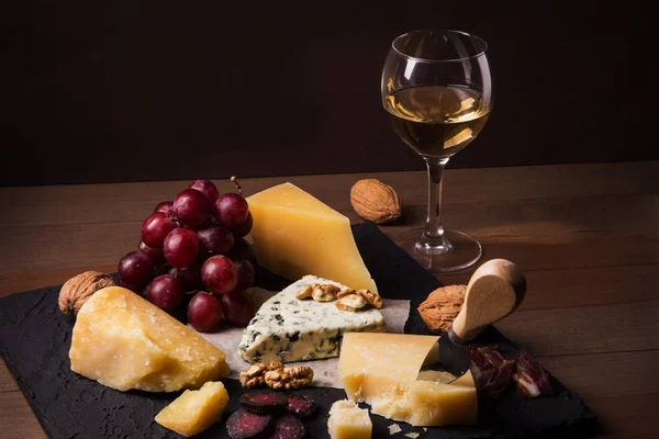 Formaggi assortiti, noci, uva, frutta, carne affumicata e un bicchiere di vino su un tavolo da portata. Scuro e stile Moody. Spazio libero per il testo . — Foto Stock