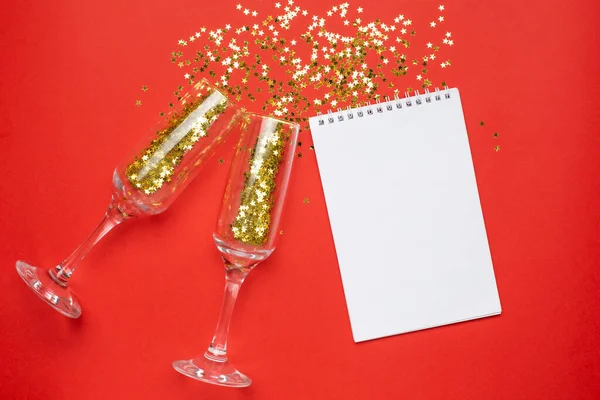 Σημειωματάριο και ποτήρια σαμπάνιας με χρυσά αστέρια κομφετί σε φόντο κόκκινο χρώμα χαρτί minimal στυλ, Χριστούγεννα και το νέο έτος έννοια — Φωτογραφία Αρχείου