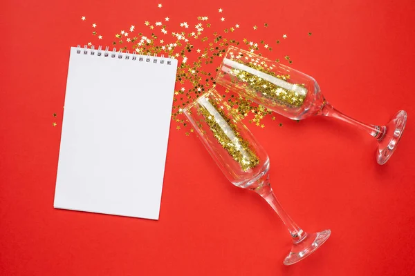 Σημειωματάριο και ποτήρια σαμπάνιας με χρυσά αστέρια κομφετί σε φόντο κόκκινο χρώμα χαρτί minimal στυλ, Χριστούγεννα και το νέο έτος έννοια — Φωτογραφία Αρχείου