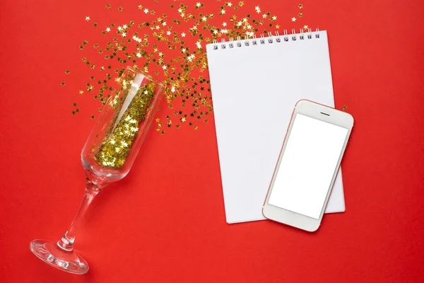 Handy, Notizblock und Sektgläser mit goldenem Sternenkonfetti auf rotem Papier Hintergrund minimaler Stil, Weihnachts- und Neujahrskonzept — Stockfoto
