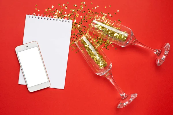 Handy, Notizblock und Sektgläser mit goldenem Sternenkonfetti auf rotem Papier Hintergrund minimaler Stil, Weihnachts- und Neujahrskonzept — Stockfoto