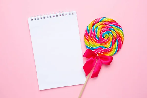 Lolipop colorat cu notebook, spirală roz, galben și alb pe fundal roz, dulciuri din copilărie — Fotografie, imagine de stoc