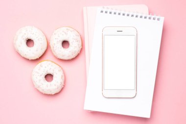 Pembe arka planda cep telefonu, defter ve çörekler.