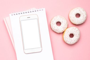 Pembe arka planda cep telefonu, defter ve çörekler.