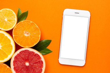 Turuncu arka planda cep telefonu ve sitruslar, meyve bezi, greyfurt, limon, mandalina ve portakallı yaz minimal kompozitonu