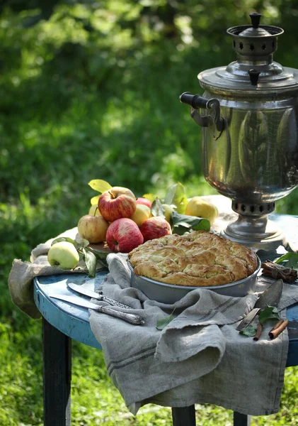 Concept of Russian cuisine. Apple biscuit cake with cinnamon. Samovar, apples, knife on an old table in the garden. Background image, copy space