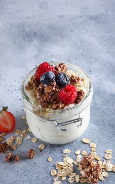 Sağlıklı kahvaltı. Şeffaf bir kavanozda yoğurt, Herkül tanecikleri, granola, taze ahududu, çilek ve bal ile doldurulmuş yaban mersini. Masadaki bal çubuğu. Arkaplan resmi, alanı kopyala