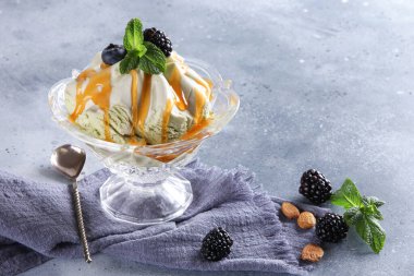 Tatlılar. Kayısı soslu dondurma ve taze böğürtlen, yaban mersini ve açık gri arka planda şeffaf bir kasede naneli şeker. Mavi kumaş, kaşık ve fındık. Arkaplan resmi, alanı kopyala