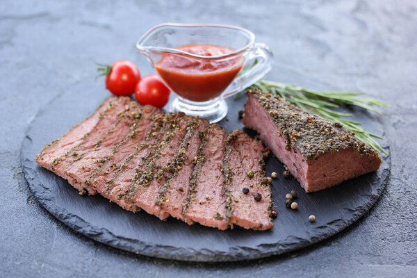 Grilled beef pastrami with rosemary, spices, cherry tomatoes and ketchup in a bowl on a black board on a dark background. Fried meat. Background image, copy space