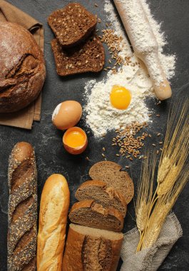 Fırın. Çeşit çeşit siyah beyaz ekmek. Tahıllı ekmek, haşhaş tohumu, susam tohumu ve baget. Buğday tohumları, çavdar kulakları, taze yumurta, un ve çuval bezi siyah arka planda.