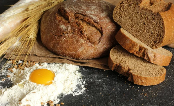 Bakery. Assortment black and white bread. Bread with cereals, poppy seeds, sesame seeds and baguettes. Wheat seeds, rye ears, fresh egg, flour and burlap on a black background
