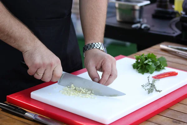 Preparation. Cooking process. The chef cuts garlic with a knife on a white board on wooden table. Thyme, red chili, coriander, herbs, chef hands. Background image, copy space