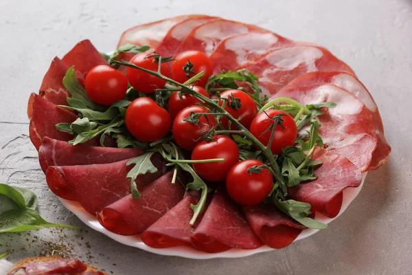 The concept of Italian cuisine. Meat snack. Bresaola and coppa with tomatoes and arugula on a white plate.