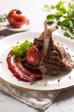 Izgara pirzola, domates ve sosla birlikte beyaz bir tabakta hafif bir arka planda. Bir demet taze kişniş, arka planda ızgara domates. Menü restoranı. Arkaplan resmi, alanı kopyala