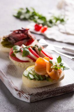 İtalyan mutfağı. Bruschetta 'nın çeşitleri. Kırmızı balıklı ekmek, avokado, caprese ve beyaz tahta üzerinde et. Açık gri arka planda domatesler ve taze otlar. Boşluğu kopyala