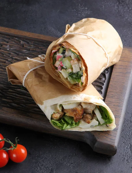 Caesar wrap. Chicken, lettuce, egg, sauce and salad in pita bread on dark wooden board on the black table. Rolls in parchment, cherry tomatoes. Shawarma vertical. Background image, copy space