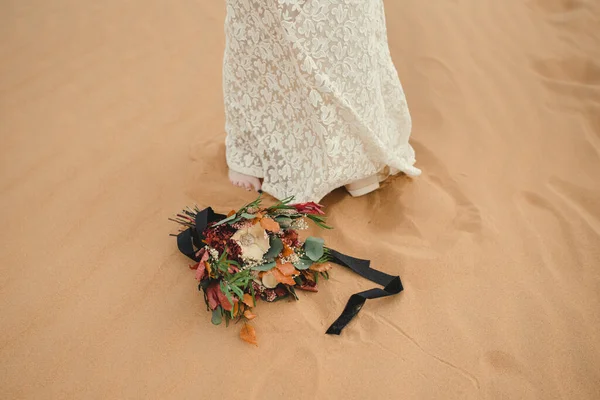 花嫁の足の隣の砂の上に横たわるブライダルブーケ. — ストック写真