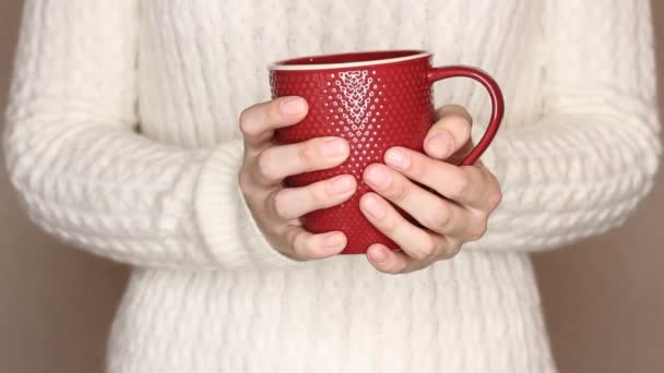 白いセーターの若い女性はお茶やコーヒーの赤いカップを保持しています 熱い飲み物で手を暖めようとして手を変える — ストック動画