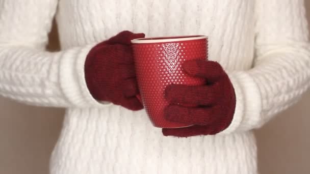 白いセーターの若い女性はお茶やコーヒーの赤いカップを保持しています 熱い飲み物で手を暖めようとして手を変える — ストック動画