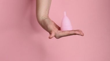 Pembe arka planda tekrar kullanılabilir pembe silikon adet bardağını tutan genç bir kadının ellerini yakın plan. Sıfır atık adet hijyen konsepti.
