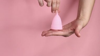 Pembe arka planda tekrar kullanılabilir pembe silikon adet bardağını tutan genç bir kadının ellerini yakın plan. Sıfır atık adet hijyen konsepti.