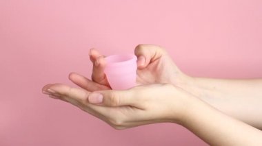 Pembe arka planda tekrar kullanılabilir pembe silikon adet bardağını tutan genç bir kadının ellerini yakın plan. Sıfır atık adet hijyen konsepti.