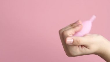 Pembe arka planda tekrar kullanılabilir pembe silikon adet bardağını tutan genç bir kadının ellerini yakın plan. Sıfır atık adet hijyen konsepti.