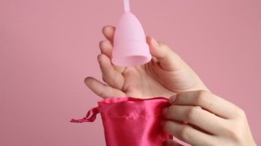 Pembe arka planda tekrar kullanılabilir pembe silikon adet bardağını tutan genç bir kadının ellerini yakın plan. Sıfır atık adet hijyen konsepti.