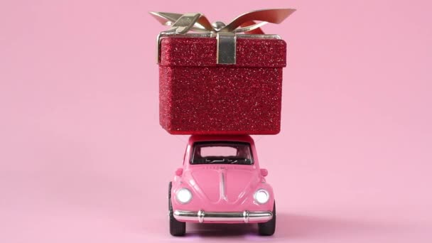 モスクワ、ロシア- 12月5 、 2019:ピンクのレトロなおもちゃの車ピンクの背景に赤いギフトボックスを提供します。お正月バレンタインデーのコンセプト — ストック動画