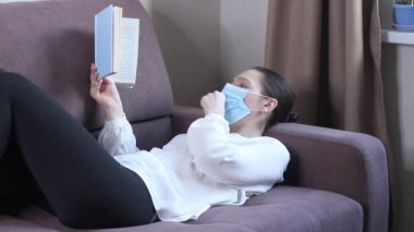 Koruyucu maskeli genç bir kadın kitap okuyor. Koronavirüs salgını. Kendini karantinaya almak için evde kal.
