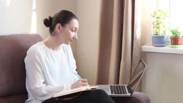 Genç kadın internet dersini ya da web sitesini dinliyor ve notlar yazıyor. Koronavirüs salgını. Kendini karantinaya almak için evde kal. — Stok video