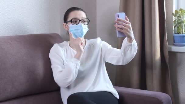 保護顔マスクを身に着けている若い女性ブロガーは 携帯電話でビデオを記録しています 女性ブロガーはコロナウイルスのパンデミックについてストリーミング 社会的距離封じ込めのための家にいる自己隔離 — ストック動画